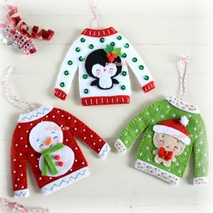 May include: Three felt jumper ornaments featuring festive designs. One white jumper with a penguin, one red jumper with a snowman, and one green jumper with a gingerbread person. Each ornament includes a hanging string and decorative accents.