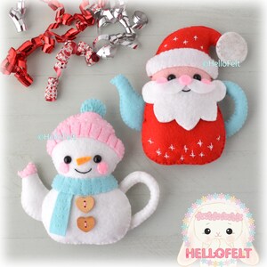 Può includere: Due teiere in feltro a tema natalizio: un pupazzo di neve con cappello rosa e sciarpa blu, e un Babbo Natale con cappello e barba rossi. Entrambe hanno manici e beccucci blu. Nastri decorativi rossi e argentati sullo sfondo.