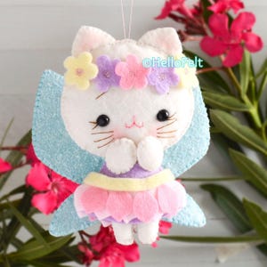 May include: A white felt cat ornament featuring a flower crown with yellow, pink, and purple blossoms. The cat has black bead eyes, stitched whiskers, and a pink skirt. It includes light blue glittery wings and a string for hanging.