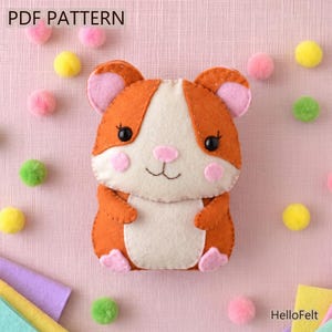 Może przedstawiać: Projekt rękodzieła z filcu przedstawiający chomika, oznaczony jako PDF PATTERN i HelloFelt. Chomik jest pomarańczowo-kremowy z różowymi uszami, policzkami i łapkami, ma czarne koralikowe oczy oraz wyszywany nos i buzię, otoczony pomponami.