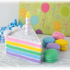 Puede incluir: Un juego de juguetes de fieltro que incluye una porción triangular de pastel arcoíris con glaseado blanco, chispas de colores y una vela, acompañado de varios macarons de fieltro pequeños, redondos y multicolores en azul, rosa, amarillo, morado y verde.