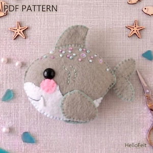 Pode incluir: Um tubarão de feltro feito à mão, cinza, branco e rosa. O tubarão tem um olho preto, boca rosa e contas decorativas. A imagem inclui o texto "PDF PATTERN" e "HelloFelt".