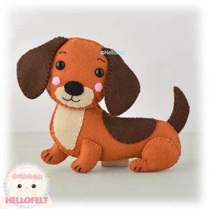 Può includere: Un giocattolo fatto a mano a forma di cane bassotto in feltro marrone, beige e nero. Il cane ha grandi orecchie flosce, un naso nero e guance rosa. Il logo "HelloFelt" è visibile.