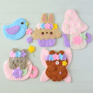 Può includere: Cinque applicazioni in feltro che includono un uccellino blu con un ramo fiorito, un cupcake a forma di coniglio beige con corona di fiori, una teiera a fungo rosa, un gatto grigio in una tazza rosa e un orso marrone con ali rosa.