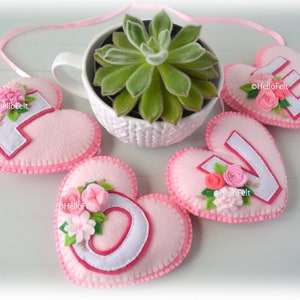 May include: Pink felt hearts with white felt letters spelling "LOVE" with pink felt flowers and green felt leaves. The hearts are strung together with a pink ribbon.