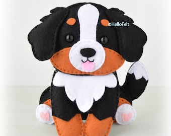 May include: A felt Bernese Mountain Dog toy with black, white, and brown fur. The dog is sitting with its paws tucked under its body. It has a pink tongue sticking out and black eyes. The text "@HelloFelt" is in the upper right corner of the image.