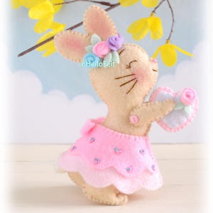 Op de afbeelding: Een beige vilt konijnfiguur met een roze rokje met kleine blauwe accenten. Het konijn heeft een bloemenkrans van blauwe, roze en groene viltbloemen op het hoofd en houdt een hartvormig ornament met een roze roos vast. De tekst HelloFelt is zichtbaar.