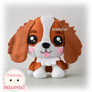 May include: A felt dog toy in brown, white, and pink. The toy dog has large black eyes, pink cheeks, and a small pink tongue. The ears are brown, and the dog has a white chest and paws. The toy is a Cavalier King Charles Spaniel.