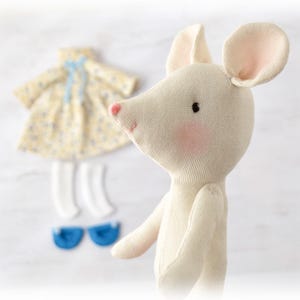 May include: A cream-coloured fabric mouse doll with pink-tinted cheeks and a small pink nose. Beside the doll is a set of clothing including a yellow floral dress with a blue ribbon, a pair of white socks, and a pair of small blue shoes.