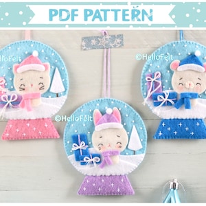 Puede incluir: Tres adornos de fieltro con forma de bola de nieve con un gato blanco, un oso blanco y un conejito blanco en su interior. Cada adorno tiene un fondo azul con copos de nieve blancos y una base blanca con un borde de color diferente. Los adornos están colgados de una cuerda y el texto "HelloFelt" es visible en cada adorno.