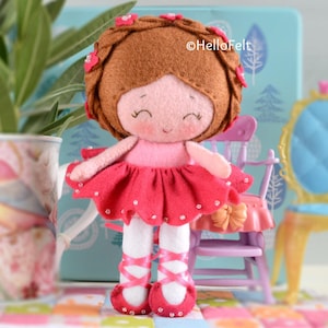 May include: A felt doll with brown hair styled in braids adorned with small pink flowers, a light pink bodice, a dark pink ruffled skirt with white bead accents, and white legs with dark pink ribbon-style ballet slippers. Text reads HelloFelt.