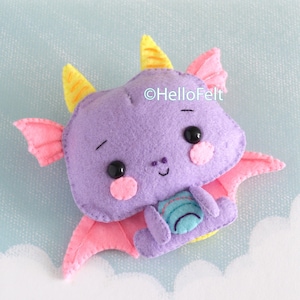 Puede incluir: Un peluche de dragón de fieltro morado con dos cuernos amarillos, alas rosas, ojos de cuentas negras y mejillas circulares rosas. El dragón tiene un pequeño parche azul en la barriga con detalles cosidos y el texto HelloFelt impreso en blanco.