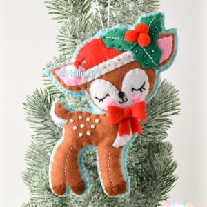May include: A felt deer ornament featuring a brown body with white spots, a red Santa hat, and a green holly leaf accent with three red berries. The deer has closed eyes, pink cheeks, and a red bow at the neck. The text HelloFelt is visible.