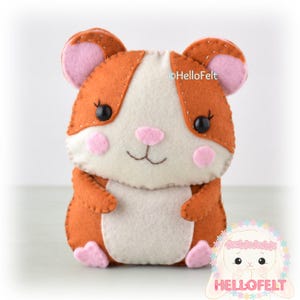 May include: A handcrafted felt hamster plush toy featuring orange and cream-colored fabric with visible white stitching. The hamster has pink ears, pink cheeks, a pink nose, and black bead eyes. The text HelloFelt is printed on the toy.