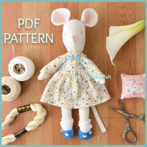 Può includere: Una bambola a forma di topo bianco fatta a mano con un vestito floreale con dettagli blu e scarpe di feltro blu. La bambola ha grandi orecchie rosa e un naso rosa. L'immagine include rocchetti di filo, filo da ricamo, un cuscinetto per spilli e forbici. Il testo sull'immagine recita "PDF PATTERN".