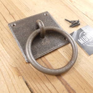 May include: A dark gray metal ring pull handle attached to a square mounting plate with four corner screw holes. The hardware features a textured finish and includes three matching black screws and a small square tag labeled MIDWEST.
