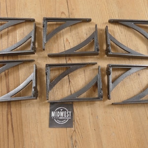 May include: Six dark gray metal shelf brackets arranged in two rows. Each bracket features a right-angle frame with a curved diagonal support brace. A small square black label with the white text MIDWEST is positioned below the center.