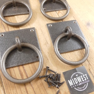 May include: Four dark gray metal ring pulls with square mounting plates, each featuring four screw holes. A small pile of matching black screws is included, along with a square card displaying the text MIDWEST CRAFT HOUSE in white.