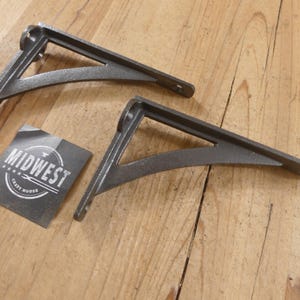 May include: Two dark gray cast iron shelf brackets with a triangular support design and pre-drilled mounting holes. A square black sticker with the white text MIDWEST CRAFT HOUSE and a graphic of a hammer is positioned between the two brackets.