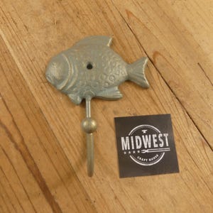 May include: A cast metal fish-shaped wall hook in a muted silver-green color. The hook has a decorative ball and a single hook. A black square with the text "MIDWEST CRAFT HOUSE" is next to the hook.