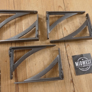 May include: Three L-shaped dark gray metal shelf brackets with a curved support brace. Each bracket features pre-drilled holes for mounting. A small square black card with the white text MIDWEST CRAFT HOUSE is positioned near the brackets.