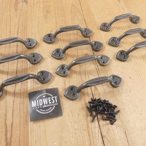 May include: A collection of ten dark gray cast iron handles with a curved design and decorative end plates. A small pile of black screws and a black and white label with the text "MIDWEST CRAFT HOUSE" are also present.