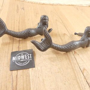 May include: Two dark gray cast iron drawer pulls shaped like mermaids with textured scales and flowing hair. Each handle features a horizontal orientation with a mounting screw visible on one. A square black label displays the text MIDWEST CRAFT HOUSE.