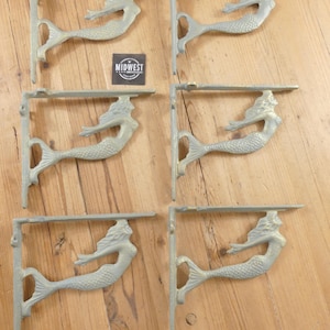 May include: Six cast iron shelf brackets with a mermaid design. The brackets are painted in a light green color and are mounted on a wooden surface. The mermaid is depicted with flowing hair and a fishtail.