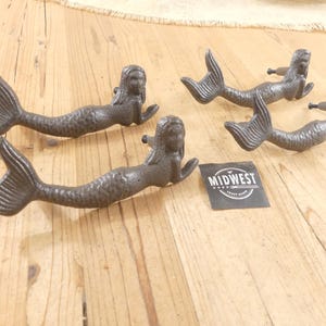 May include: Three dark gray cast iron drawer pulls shaped like mermaids with textured scales and defined tails. Each handle features a reclining mermaid figure with arms extended forward. A small black square tag with the white text MIDWEST is included.