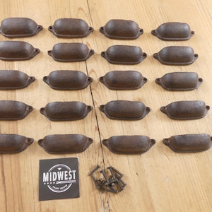 May include: A collection of twenty cast iron drawer pulls with a rustic, weathered finish. Each pull is oval-shaped with a small handle on each end. A black and white label with the text "MIDWEST CRAFT HOUSE" is also present.