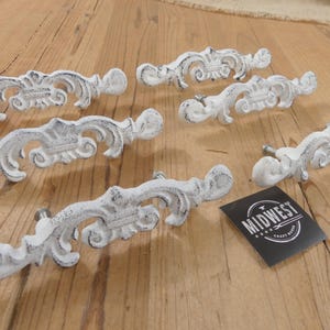 May include: White distressed decorative drawer pulls with ornate scrollwork. The hardware is arranged on a wooden surface. A black tag with the text "MIDWEST CRAFT GOODS" is also present.