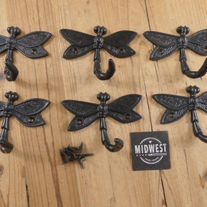 May include: Six black cast iron dragonfly hooks with curved hooks, arranged on a wooden surface. The dragonfly wings are detailed. A small pile of screws and a black square with the text "MIDWEST CRAFT HOUSE" are also present.