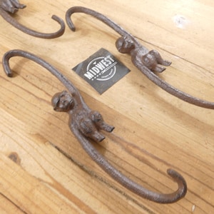 May include: Two brown, textured metal S-shaped hooks, each featuring a monkey figure integrated into the center. A small square label with the text MIDWEST CRAFT WORK is positioned between the two hooks, which are arranged diagonally.