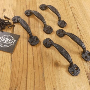 May include: Five black cast iron handles with decorative floral patterns and matching screws are arranged on a light wood surface. A black and white label with the text "MIDWEST CRAFT HOUSE" is also present.