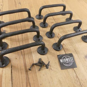 May include: A set of eight black cast iron cabinet handles with a textured finish, each featuring two circular mounting plates with pre-drilled holes. A small pile of matching black screws and a square black label with the text MIDWEST CRAFT HOUSE.