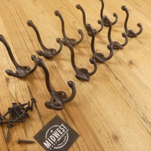 May include: Set of eight antique-style cast iron coat hooks with mounting screws. The hooks are dark brown with a curved design and a double hook. The logo for Midwest Craft House is visible.