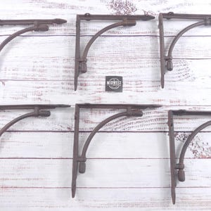 May include: Six rustic, brown metal shelf brackets. Each bracket has a curved support arm and a decorative top. The brackets are arranged on a white wooden surface. The Midwest logo is visible.