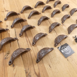 May include: A collection of twenty-one dark brown cast iron cup drawer pulls featuring an embossed scrollwork pattern. Each handle has two screw holes for mounting. A small black square card with the white text MIDWEST is included.