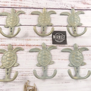 May include: Six decorative wall hooks in the shape of sea turtles. The hooks are a muted green color with an anchor-shaped hook below the turtle. The hooks are arranged on a white wooden surface. A black and white label reads "Midwest".