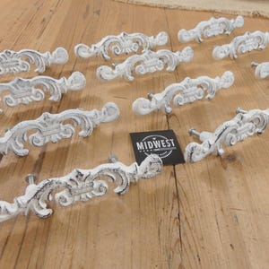 May include: A collection of white, ornate drawer pulls with a distressed finish. The pulls feature a scrolling design and are arranged on a wooden surface. A black and white label with the text "MIDWEST" is also present.