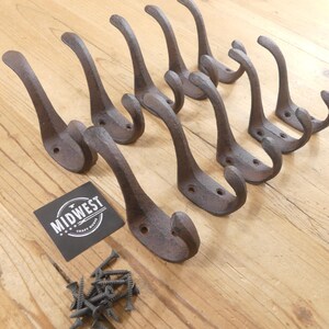 May include: A collection of eight rustic, cast iron coat hooks with a curved design. The hooks are a dark brown color and are arranged on a light wood surface. A small pile of screws and a black label with the text "MIDWEST" are also present.