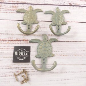 May include: Three cast iron wall hooks in the shape of turtles with anchor-shaped hooks. The hooks are painted in a muted green-gold color. A small black square with the text "MIDWEST CRAFT HOUSE" is also in the image. Several screws are also visible.