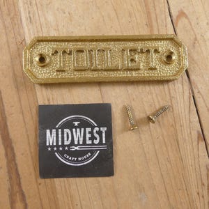 May include: A gold-colored metal sign that reads "TOILET" in embossed letters. The sign is rectangular with rounded corners and has two screw holes. A black square with the text "MIDWEST CRAFT HOUSE" and two screws are also visible.