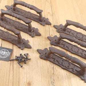 May include: Six cast iron door pulls with a rustic, brown finish. Each pull has a decorative floral design at the ends and a curved handle. A small pile of screws and a "Midwest" label are also present.