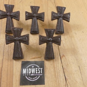 May include: Five dark brown, textured metal cross-shaped knobs arranged in a group. Below the knobs is a square black label with white text that reads MIDWEST CRAFT HOUSE, featuring a small anvil icon and four stars above the text.