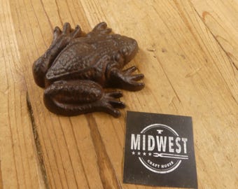 May include: A cast iron frog figurine in a dark brown finish. The frog is lying down with its legs extended. A black square with the text "MIDWEST CRAFT HOUSE" is next to the frog.