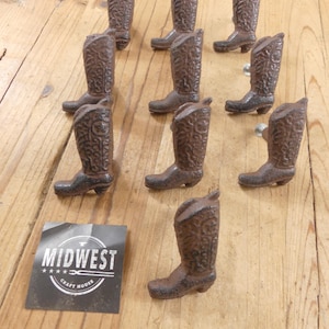 May include: A collection of ten brown cast iron cabinet knobs shaped like cowboy boots with intricate embossed floral patterns. Each boot knob features a textured finish and a metal mounting screw. A square black label displays the text MIDWEST CRAFT HOUSE.