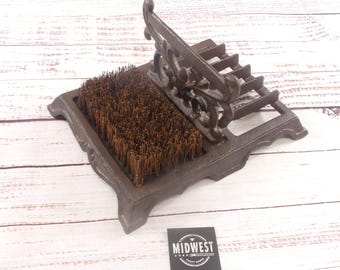 May include: A cast iron boot scraper with a built-in brush. The boot scraper has an ornate design with a rectangular base, a brush, and a decorative top. The brush is made of brown bristles. The item is brown.
