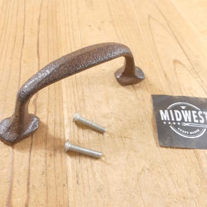 May include: A dark brown, textured cast iron handle with a curved design, accompanied by two silver-colored metal screws. A small black square label is present with the white text MIDWEST CRAFT HOUSE and a graphic of an anvil and tools.