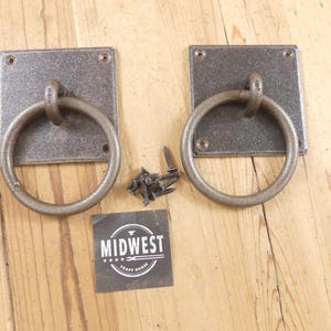 May include: Two dark gray metal ring pulls with square mounting plates, each featuring four screw holes. A small pile of matching metal screws is positioned between the pulls, alongside a black square label with the text MIDWEST CRAFT HOUSE.